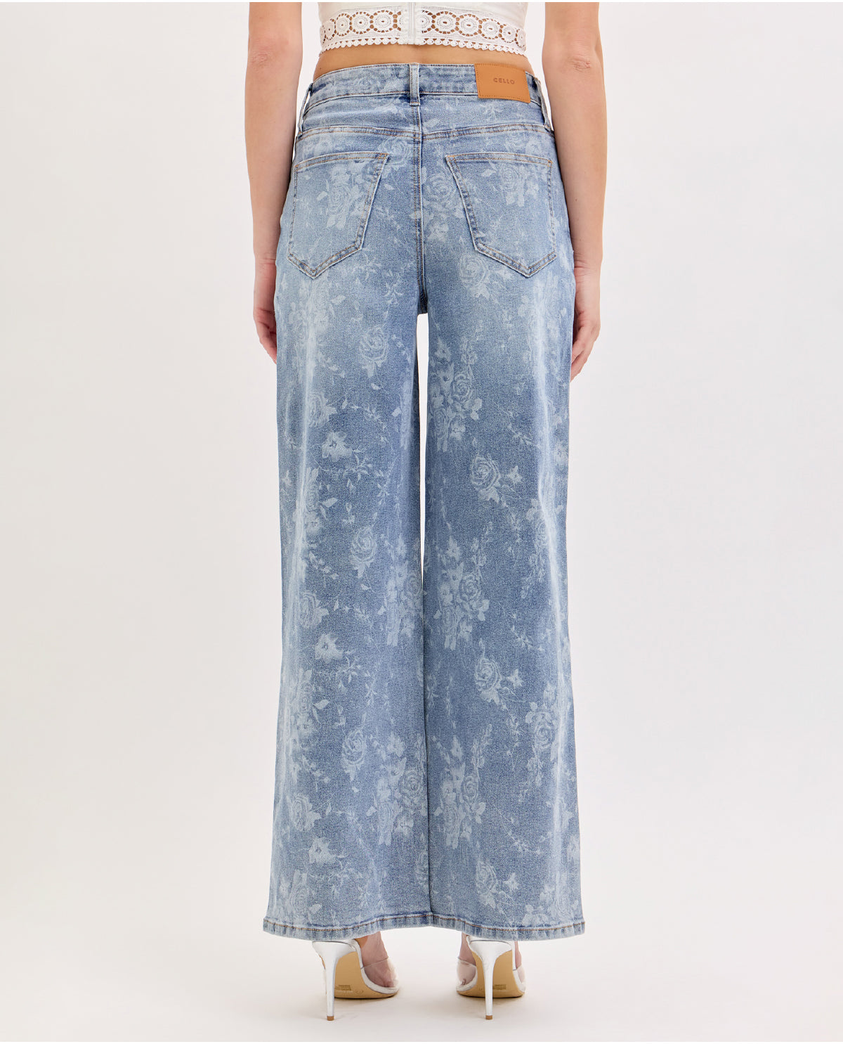 A model shown from the back wearing high-waisted, wide-leg light blue denim jeans with a white floral print and a tan leather patch on the back waistband, paired with beige high heels against a plain white studio background.