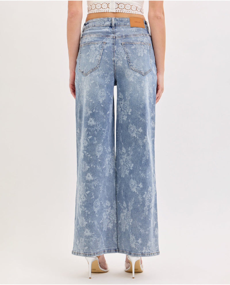 A model shown from the back wearing high-waisted, wide-leg light blue denim jeans with a white floral print and a tan leather patch on the back waistband, paired with beige high heels against a plain white studio background.