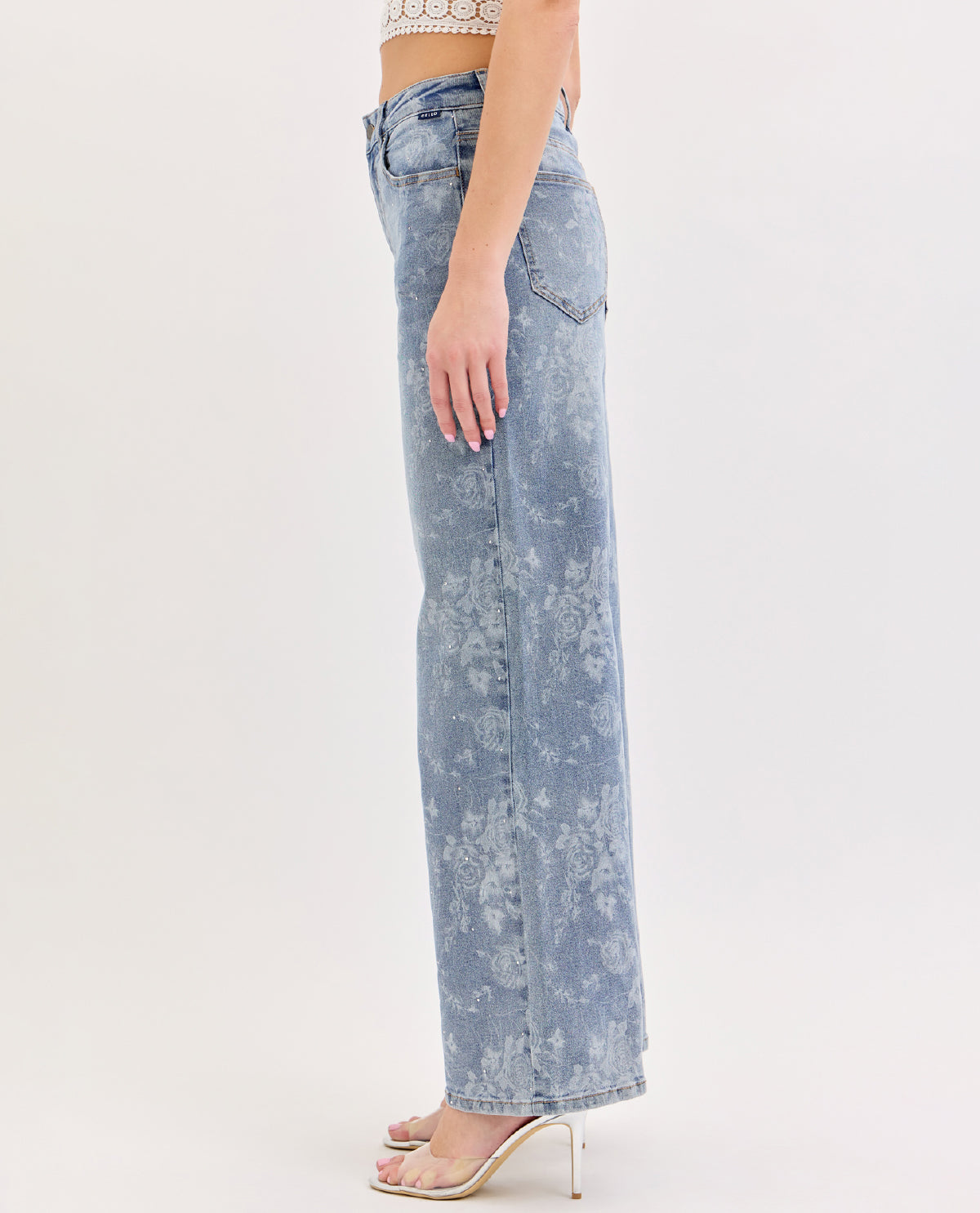 A side-profile view of a model wearing high-waisted, wide-leg light blue acid-wash jeans with a distressed mottled pattern, paired with clear high-heeled sandals against a plain white studio background.
