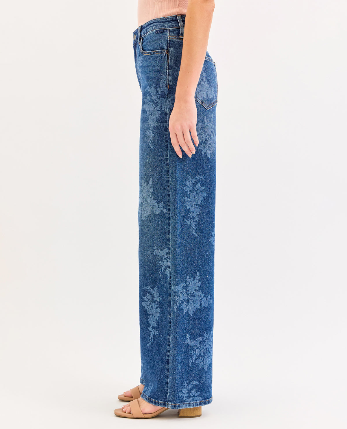 A side-profile view of a model wearing high-waisted, wide-leg blue denim jeans with a faded floral pattern, paired with tan flat sandals against a plain white studio background.