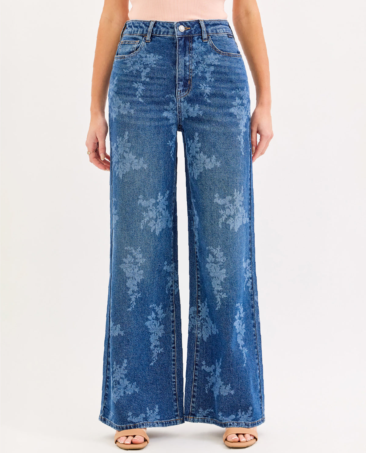 A model wearing high-waisted, wide-leg blue denim jeans with a faded floral pattern, paired with a light pink top and tan sandals, against a plain white studio background.