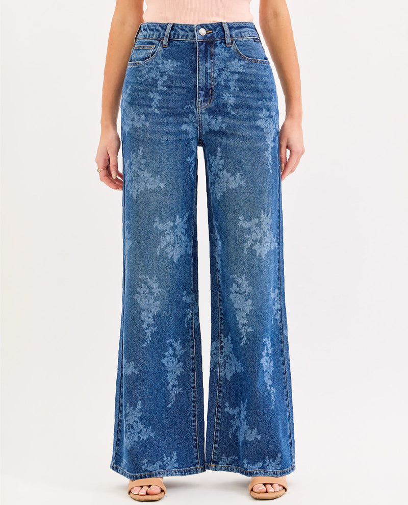 A model wearing high-waisted, wide-leg blue denim jeans with a faded floral pattern, paired with a light pink top and tan sandals, against a plain white studio background.