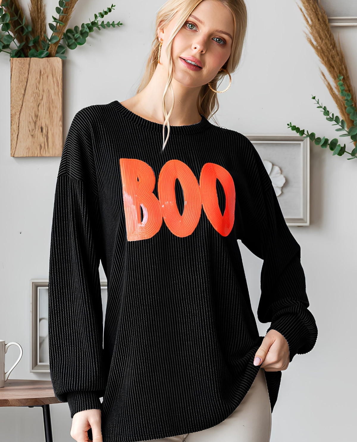 A woman wearing a black ribbed long-sleeve sweater with a large orange 'BOO' graphic across the chest, posed in a styled indoor setting with home decor elements.