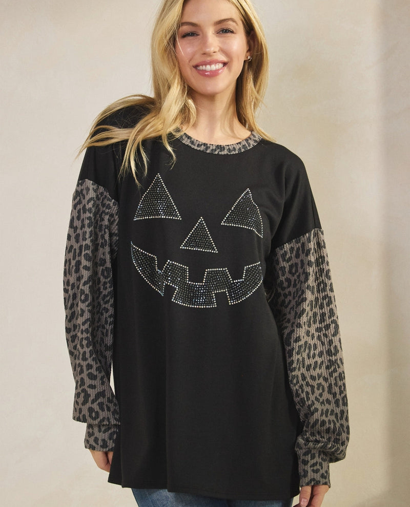A woman wearing a black long-sleeve top with leopard-print sleeves and a rhinestone jack-o'-lantern face on the front, paired with blue jeans against a neutral beige background.