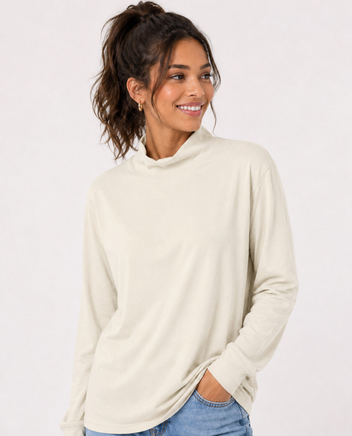 A woman wearing a cream-colored long-sleeve mock neck top and blue jeans, posing against a solid white background.