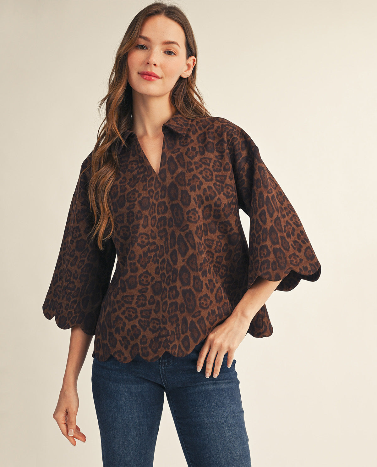 A woman modeling a brown and black leopard print blouse with wide bell sleeves and scalloped edges, paired with blue jeans, against a plain beige studio background.