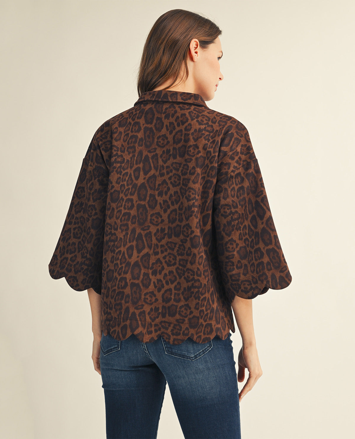 A woman shown from the back wearing a brown leopard-print short-sleeve blouse with scalloped edges and blue jeans, against a plain light beige studio background.