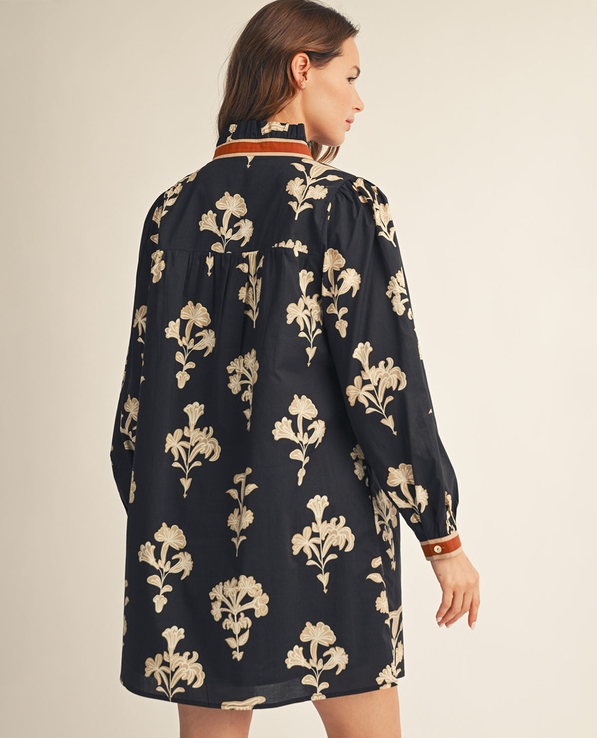 A woman shown from the back wearing a loose black long-sleeve shirt with a beige floral pattern and a striped collar, photographed against a plain light beige studio background.