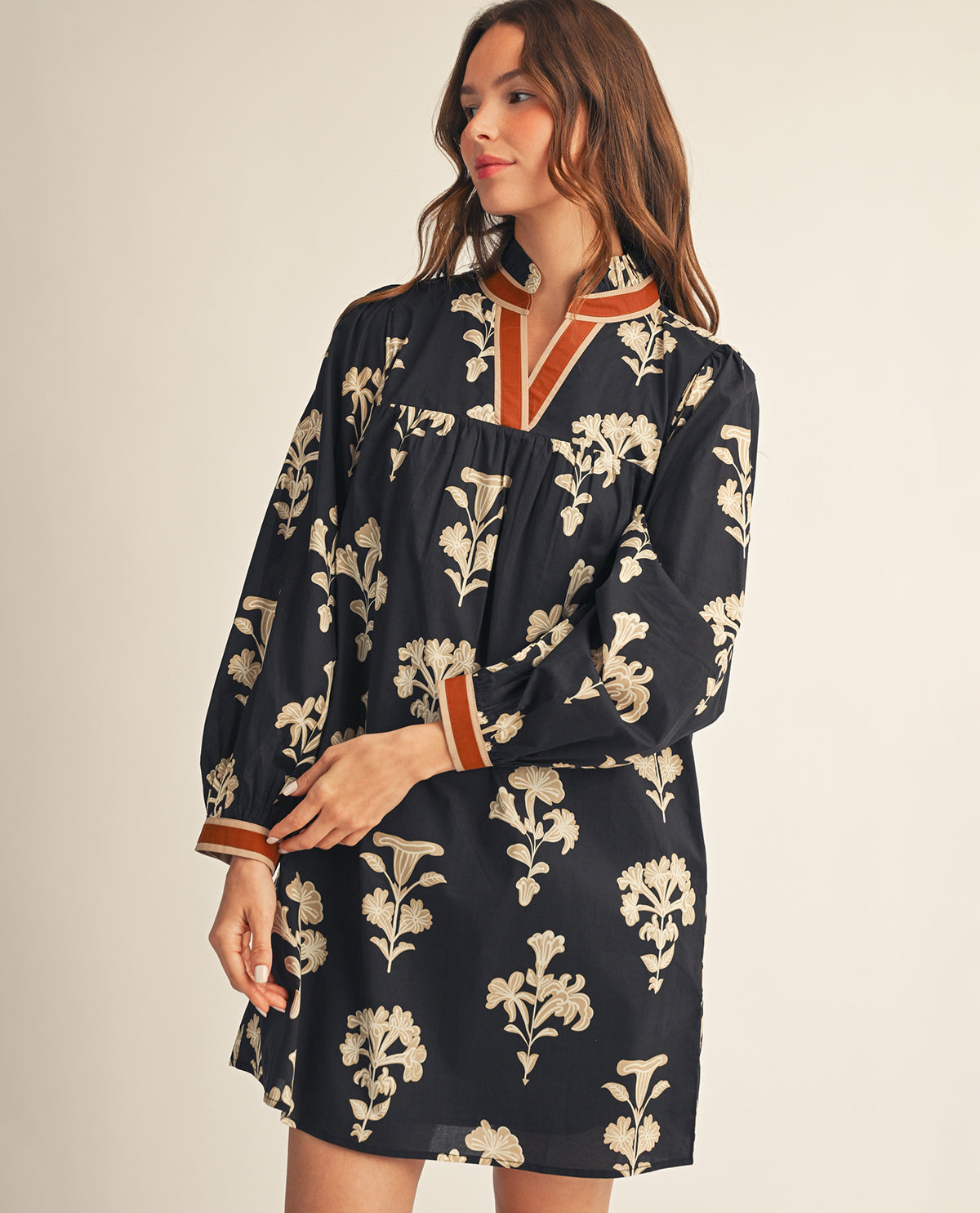 A woman wearing a long-sleeve black dress with a beige floral print and rust-orange trim on the collar and cuffs, posed against a solid light beige background.