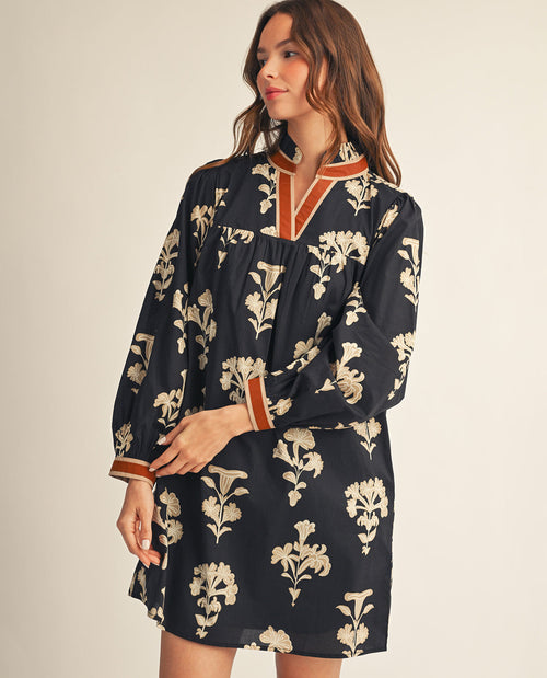 A woman wearing a long-sleeve black dress with a beige floral print and rust-orange trim on the collar and cuffs, posed against a solid light beige background.