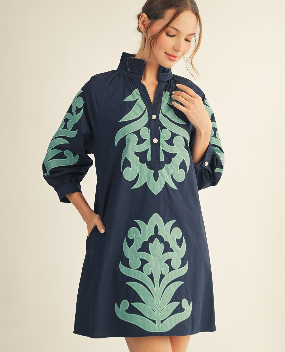 A woman modeling a navy blue shift dress with mint green embroidered patterns on the sleeves and front, set against a plain beige background.