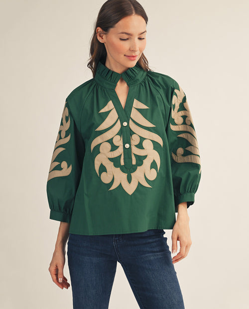 A woman wearing a dark green blouse with beige embroidered patterns on the front and sleeves, paired with dark blue jeans, against a plain beige background.