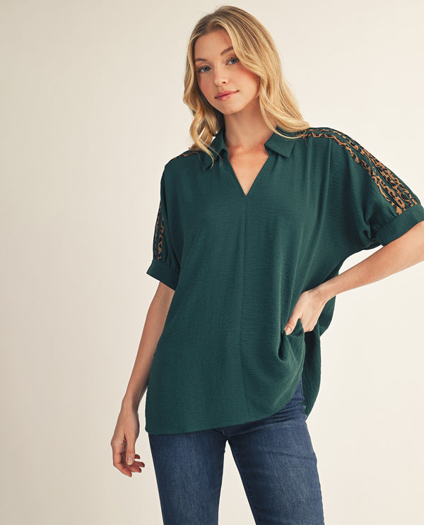 A woman modeling a dark green short-sleeve blouse with lace-up sleeves and gold patterned detailing on the shoulders, paired with blue jeans against a plain beige background.