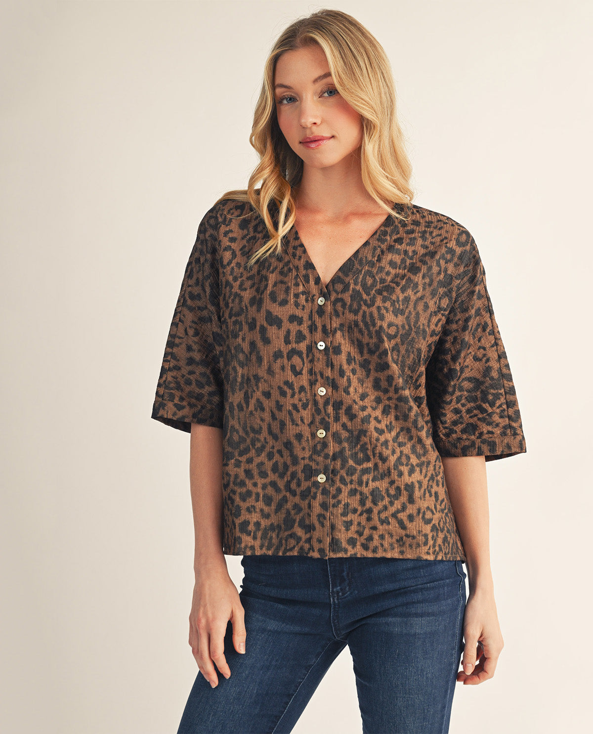 A woman modeling a brown and black leopard print short-sleeve button-up blouse with dark blue jeans, set against a plain beige studio background.