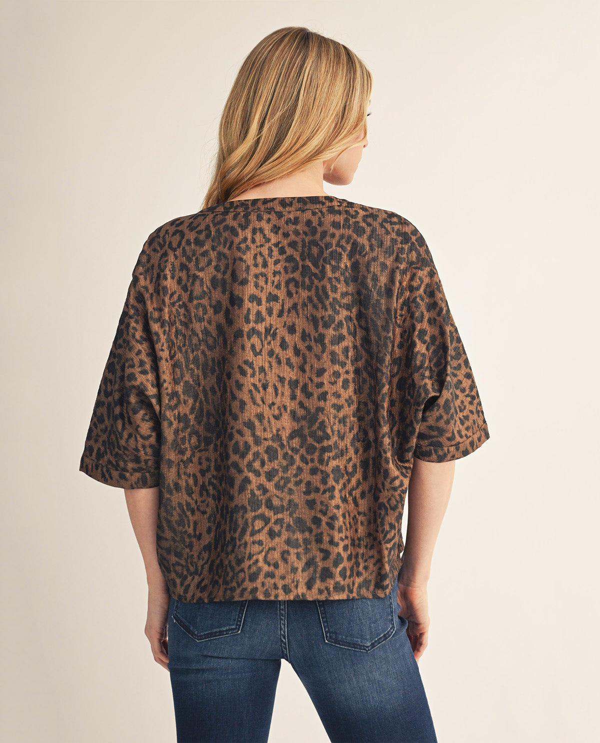 A woman viewed from behind wearing a loose-fitting leopard print top with three-quarter sleeves and blue jeans, set against a plain light beige studio background.