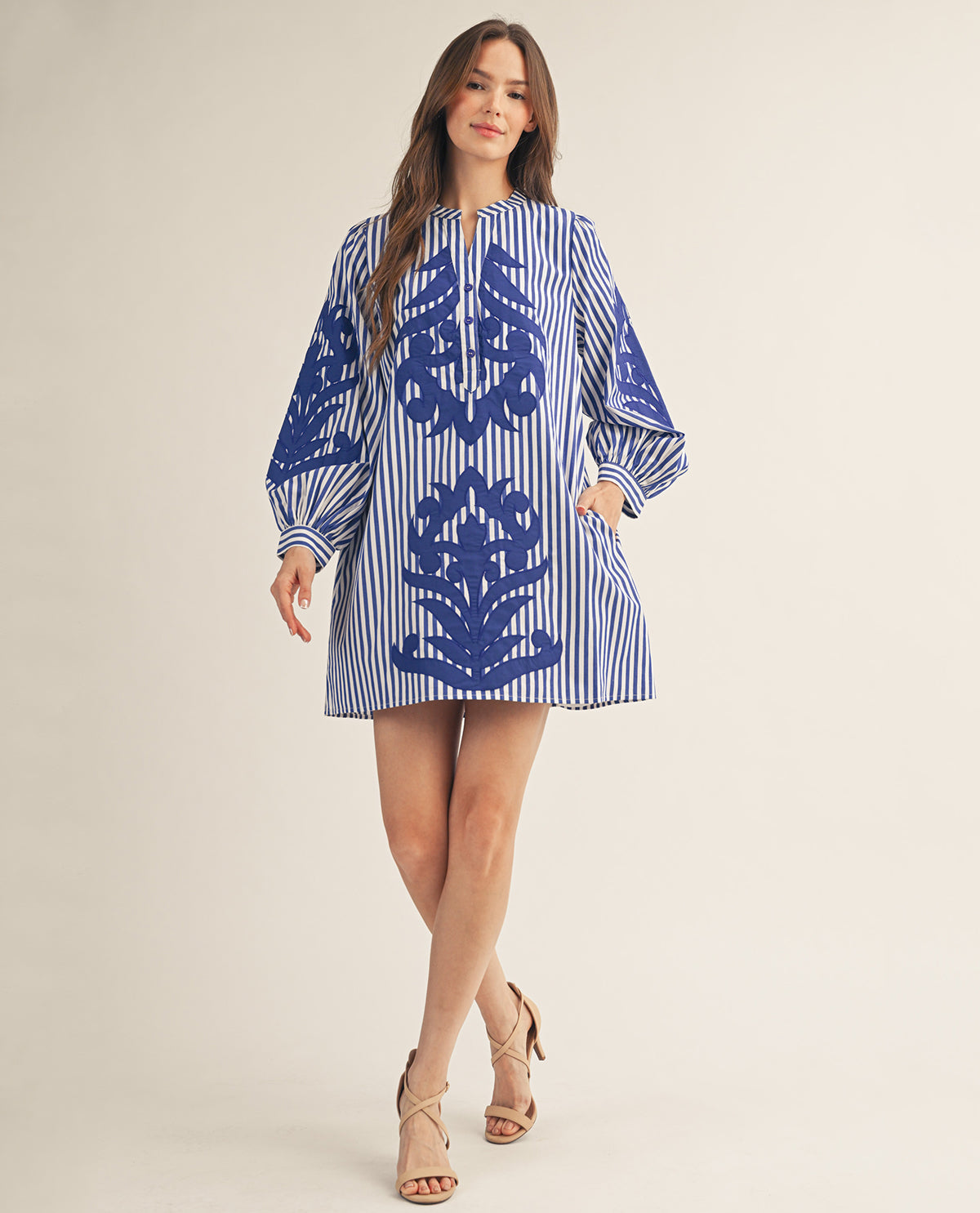 A woman modeling a blue and white striped tunic dress with a large blue floral pattern on the front, styled with beige strappy heels against a plain beige studio background.