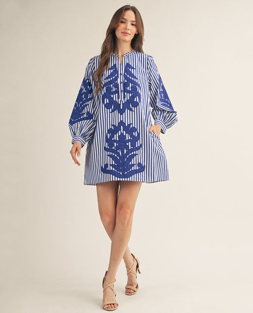A woman modeling a blue and white striped tunic dress with a large blue floral pattern on the front, styled with beige strappy heels against a plain beige studio background.