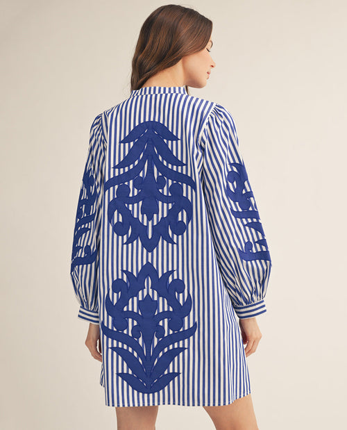 A woman shown from the back wearing a blue and white striped shirt dress with large blue floral embroidery on the back and sleeves, set against a plain beige studio background.