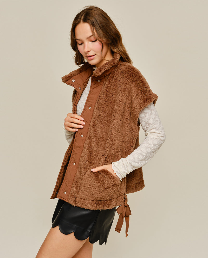 A woman models a brown sherpa-lined sleeveless button-front vest over a cream ribbed long-sleeve top and black skirt, against a solid light beige background.