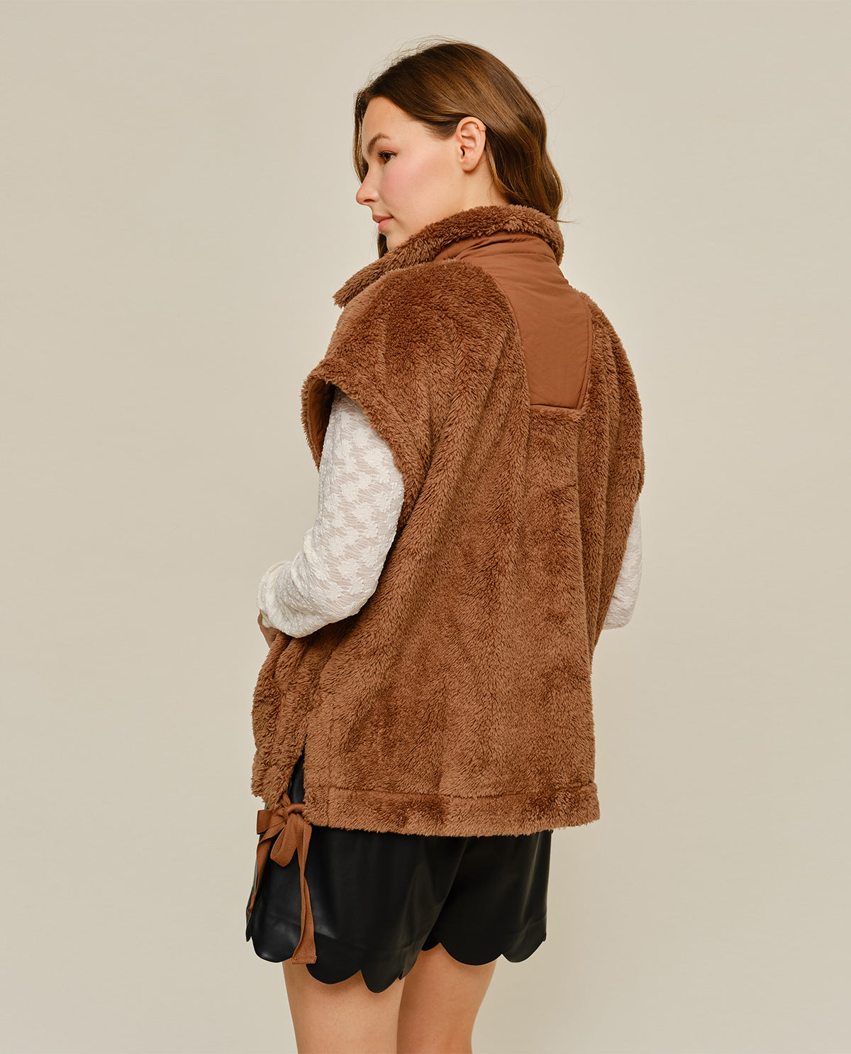 A woman shown from the back wearing a brown fuzzy sherpa vest over a cream patterned long-sleeve top and black shorts, against a plain light beige studio background.