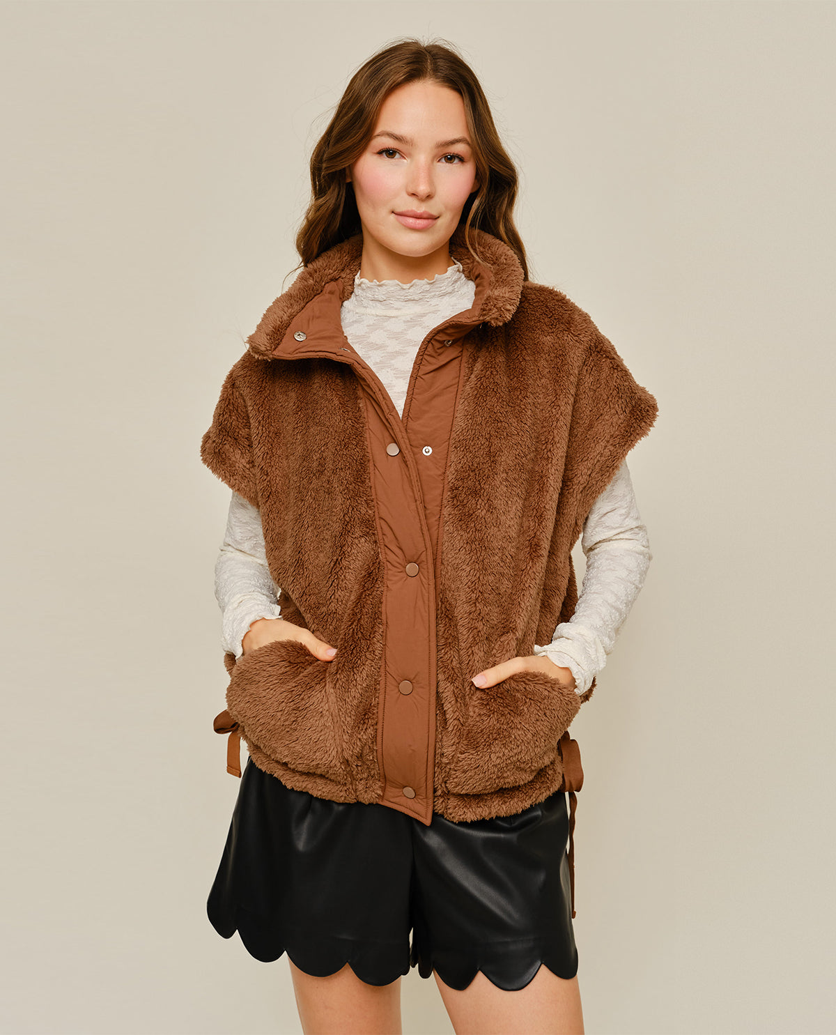 A woman modeling a brown fuzzy sleeveless vest over a cream long-sleeve top and black scalloped shorts, photographed against a plain light beige studio background.