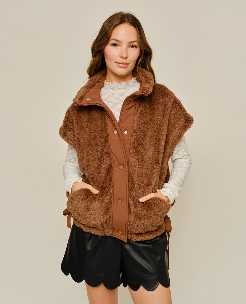 A woman modeling a brown fuzzy sleeveless vest over a cream long-sleeve top and black scalloped shorts, photographed against a plain light beige studio background.