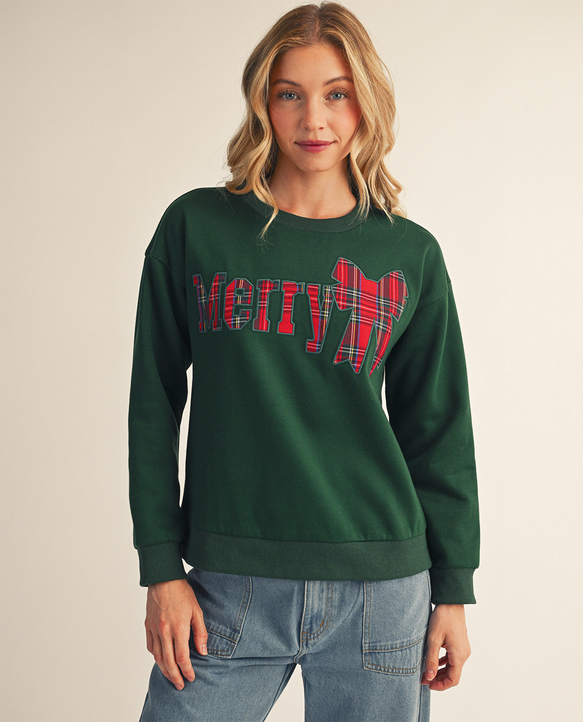 A woman wearing a dark green crewneck sweatshirt with a 'Merry' holiday graphic in red and green plaid, paired with light blue jeans, against a plain beige background.