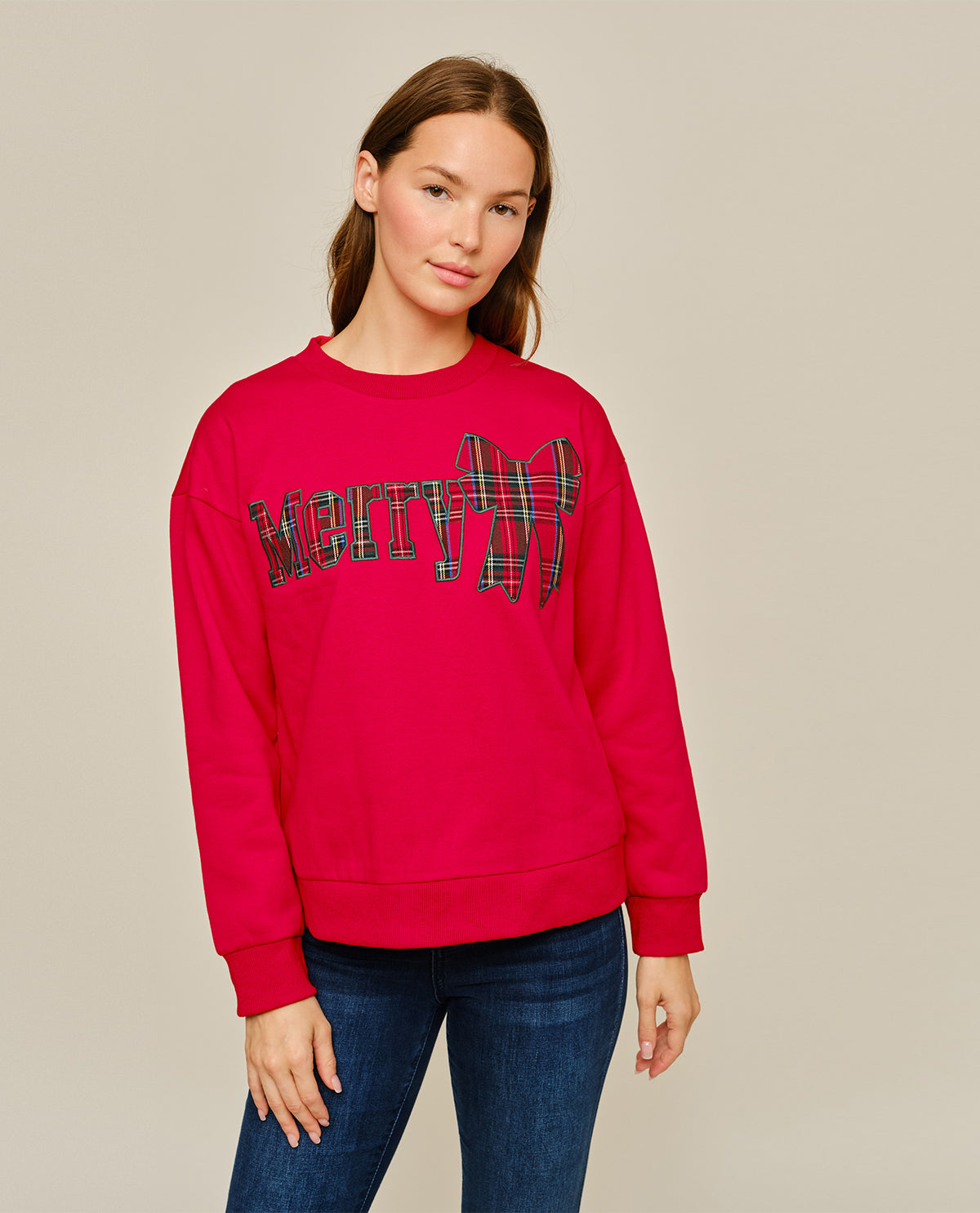A woman modeling a bright pink crewneck sweatshirt with a stylized 'MOON' graphic in a plaid pattern across the chest, paired with dark blue jeans against a solid light beige background.