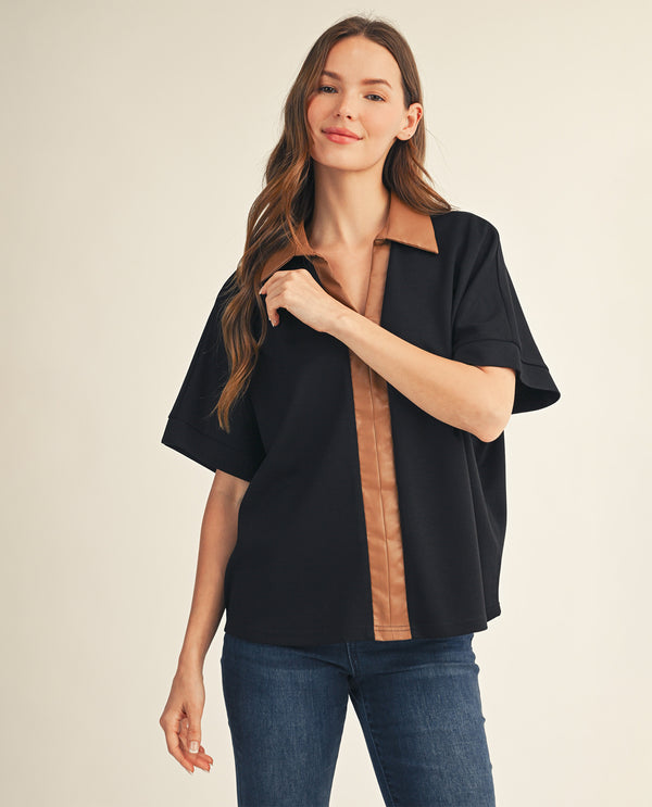 A woman modeling a black short-sleeve button-up shirt with tan faux-leather trim on the collar, front placket, and cuffs, paired with blue jeans against a warm beige studio background.