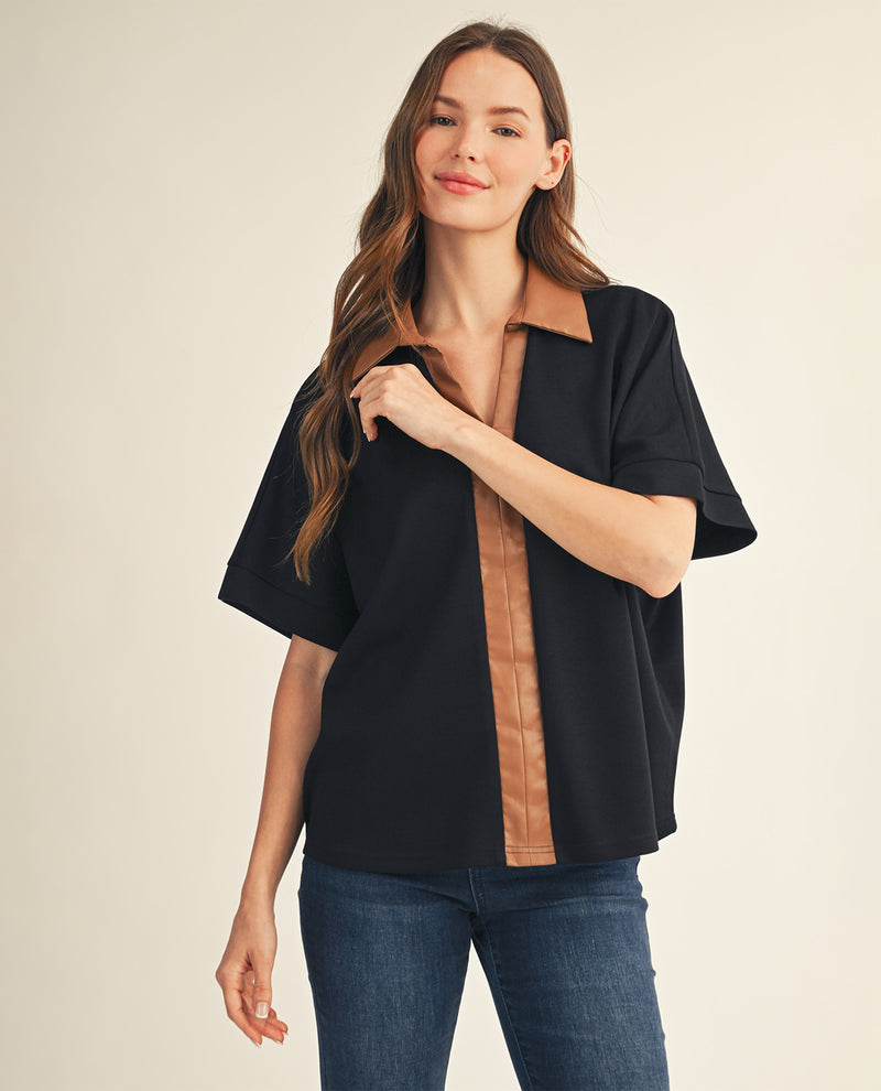 A woman modeling a black short-sleeve button-up shirt with tan faux-leather trim on the collar, front placket, and cuffs, paired with blue jeans against a warm beige studio background.