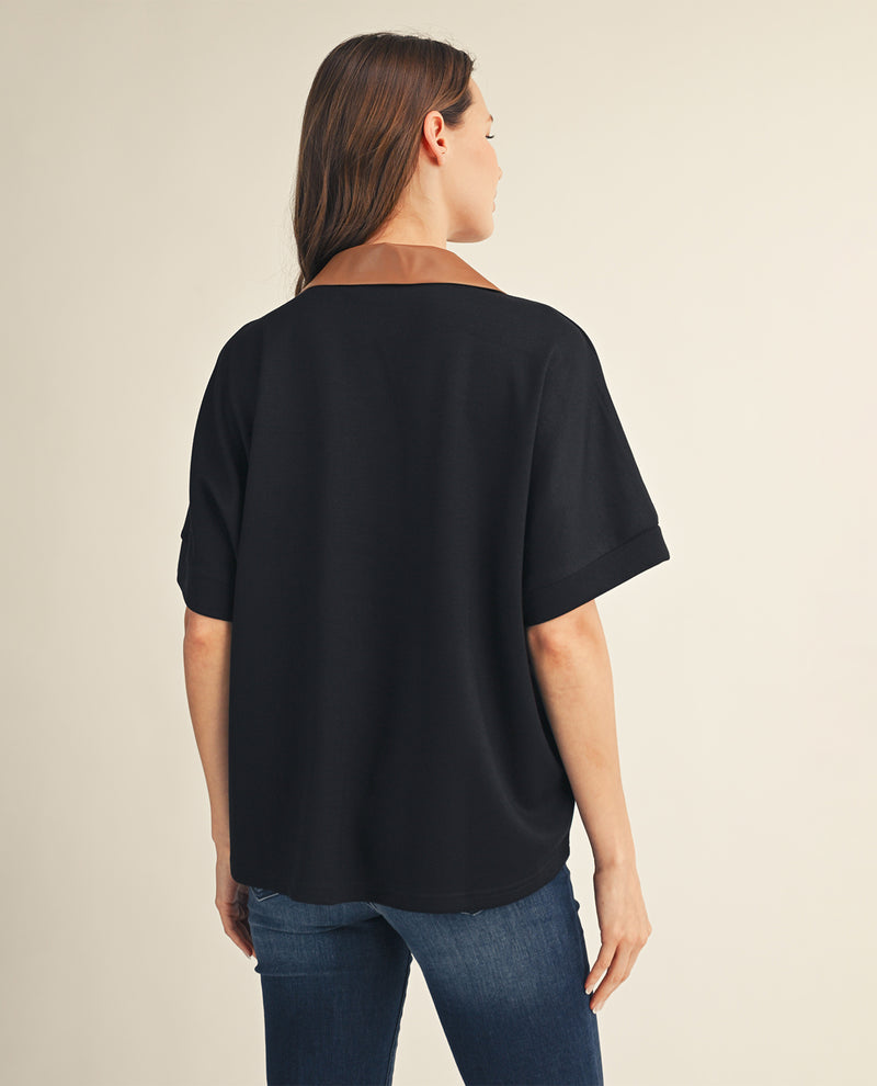 A woman viewed from the back wearing a loose black short-sleeve top with a brown collar and blue jeans, against a plain light beige studio background.