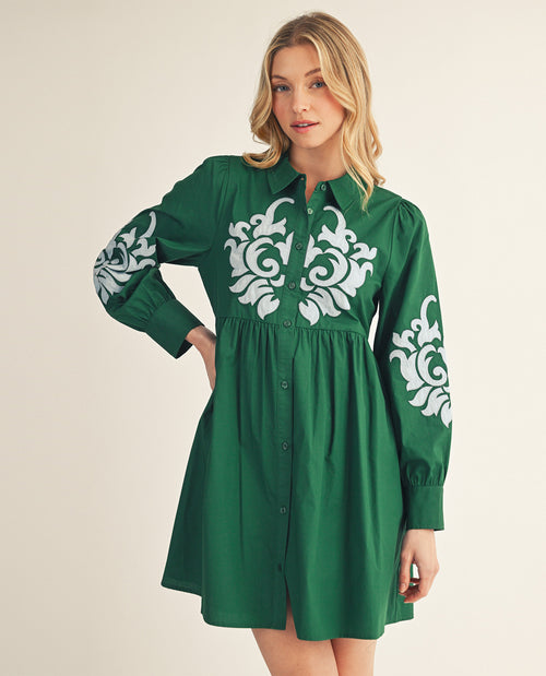 A woman modeling a green long-sleeve button-front dress with white ornate floral embroidery on the chest and sleeves, photographed against a plain light beige studio background.