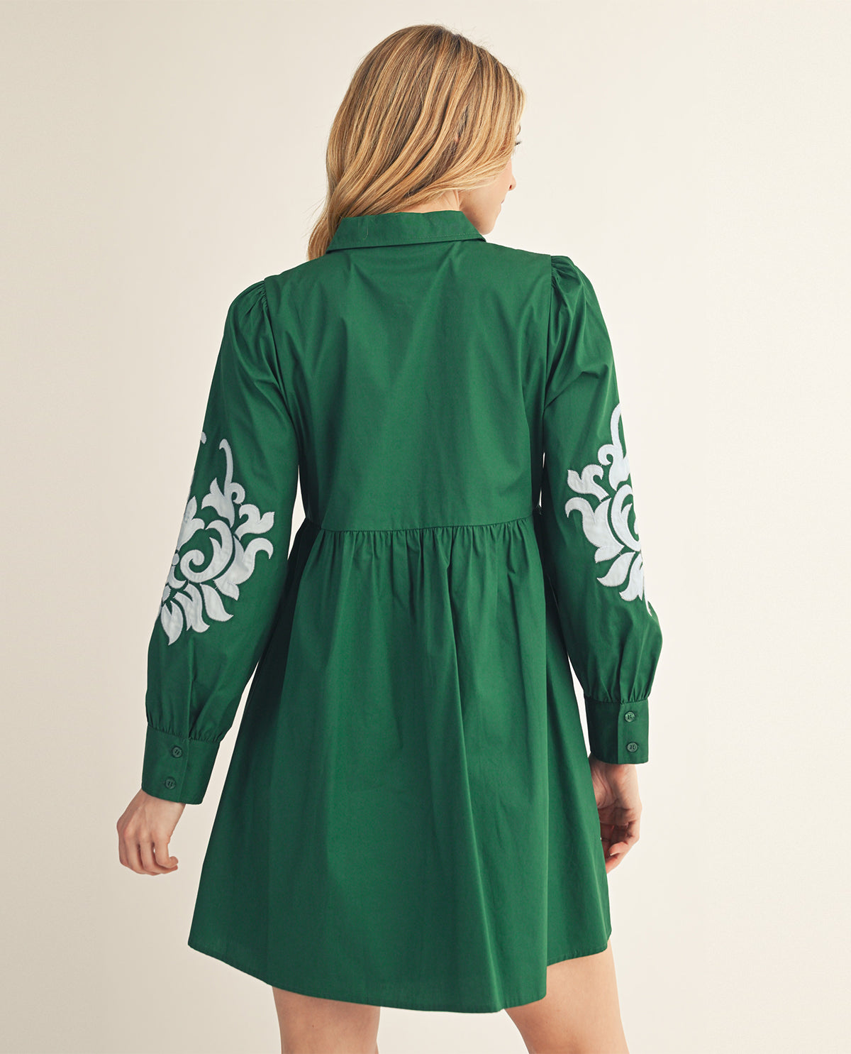 A woman shown from the back wearing a green long-sleeve mini dress with white floral embroidery on the sleeves, set against a plain light beige background.