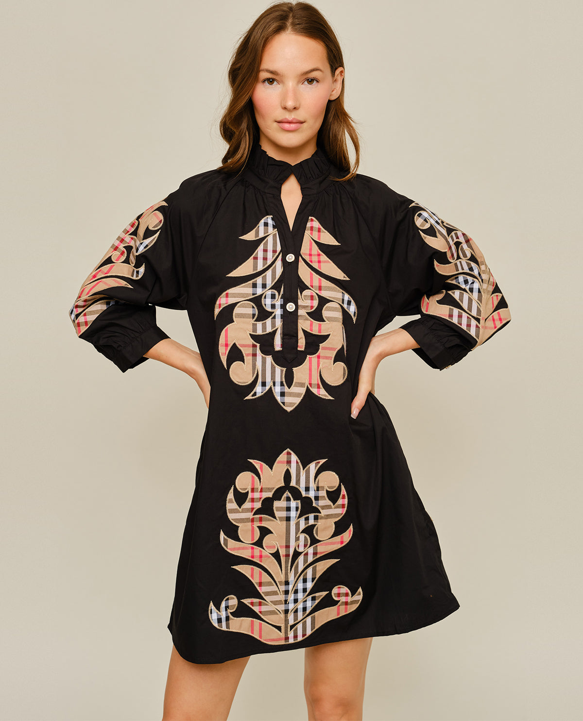 A woman modeling a black long-sleeve mini dress with a high keyhole collar and large beige, pink, and white plaid damask-style motifs on the front and sleeves, set against a solid light beige background.