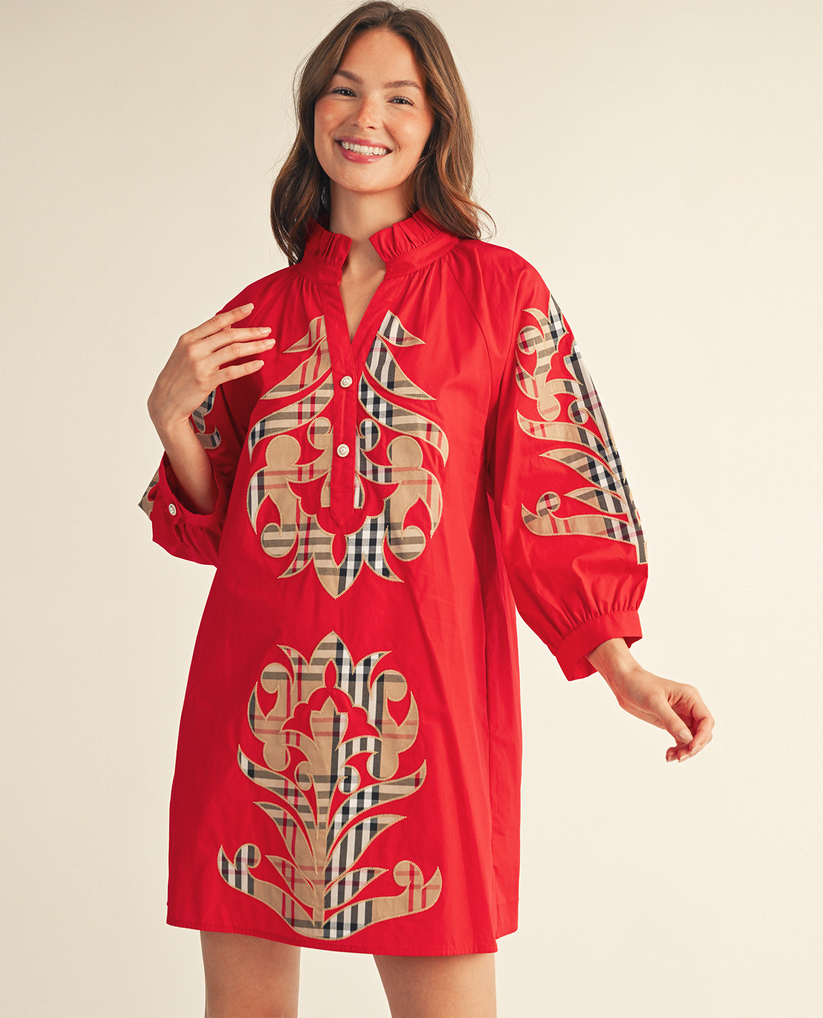 A woman modeling a red long-sleeve tunic dress with beige and black plaid patterned motifs on the front and sleeves, set against a plain beige background.