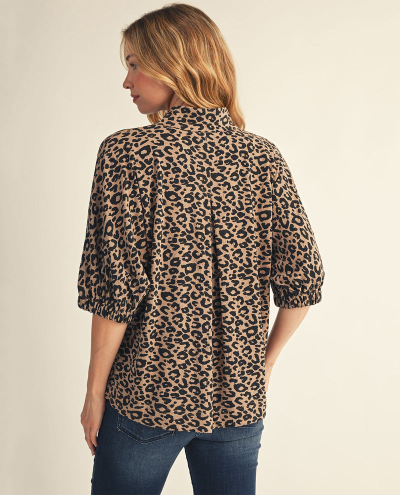 A woman shown from the back wearing a leopard-print blouse with three-quarter sleeves and blue jeans, photographed against a plain light beige studio background.