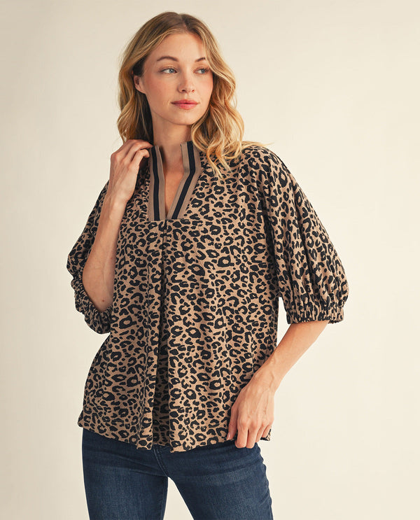 A woman wearing a leopard-print blouse with puff sleeves and a split neckline, paired with dark blue jeans, against a plain beige background.