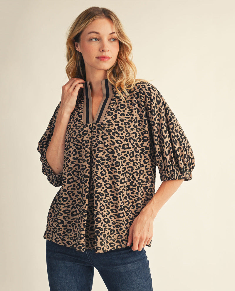 A woman wearing a leopard-print blouse with puff sleeves and a split neckline, paired with dark blue jeans, against a plain beige background.