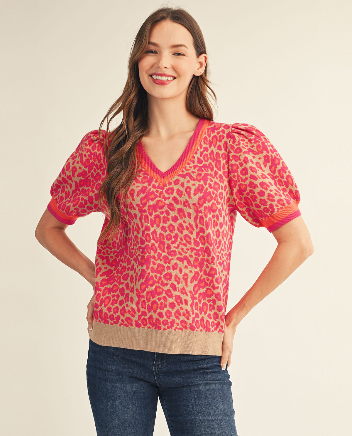 A woman modeling a short-sleeve knit top with a pink and beige leopard print, featuring puff sleeves, a V-neckline, and a ribbed hem, paired with blue jeans against a light beige studio background.