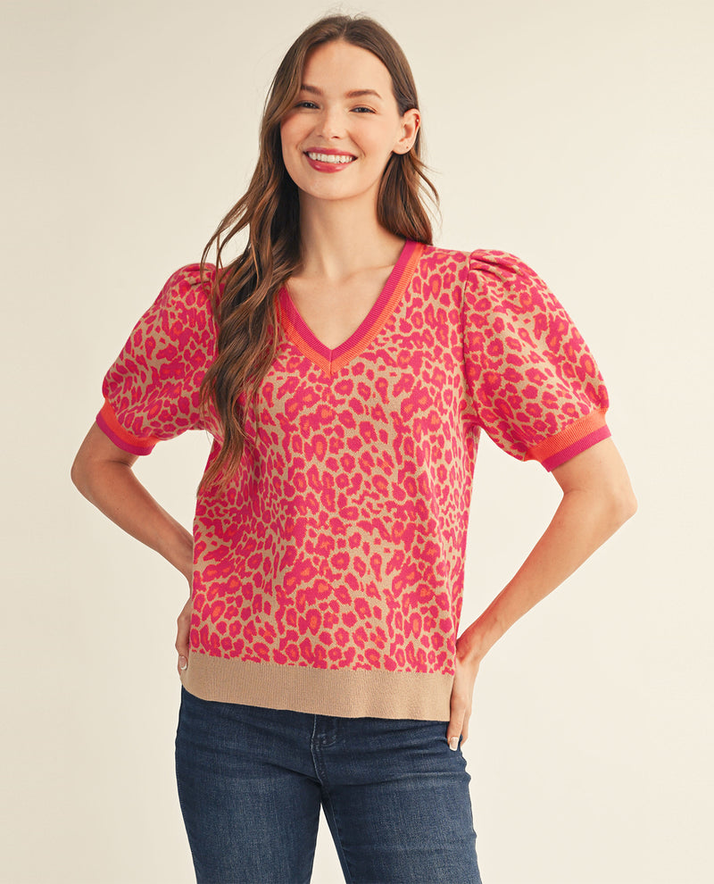 A woman modeling a short-sleeve knit top with a pink and beige leopard print, featuring puff sleeves, a V-neckline, and a ribbed hem, paired with blue jeans against a light beige studio background.