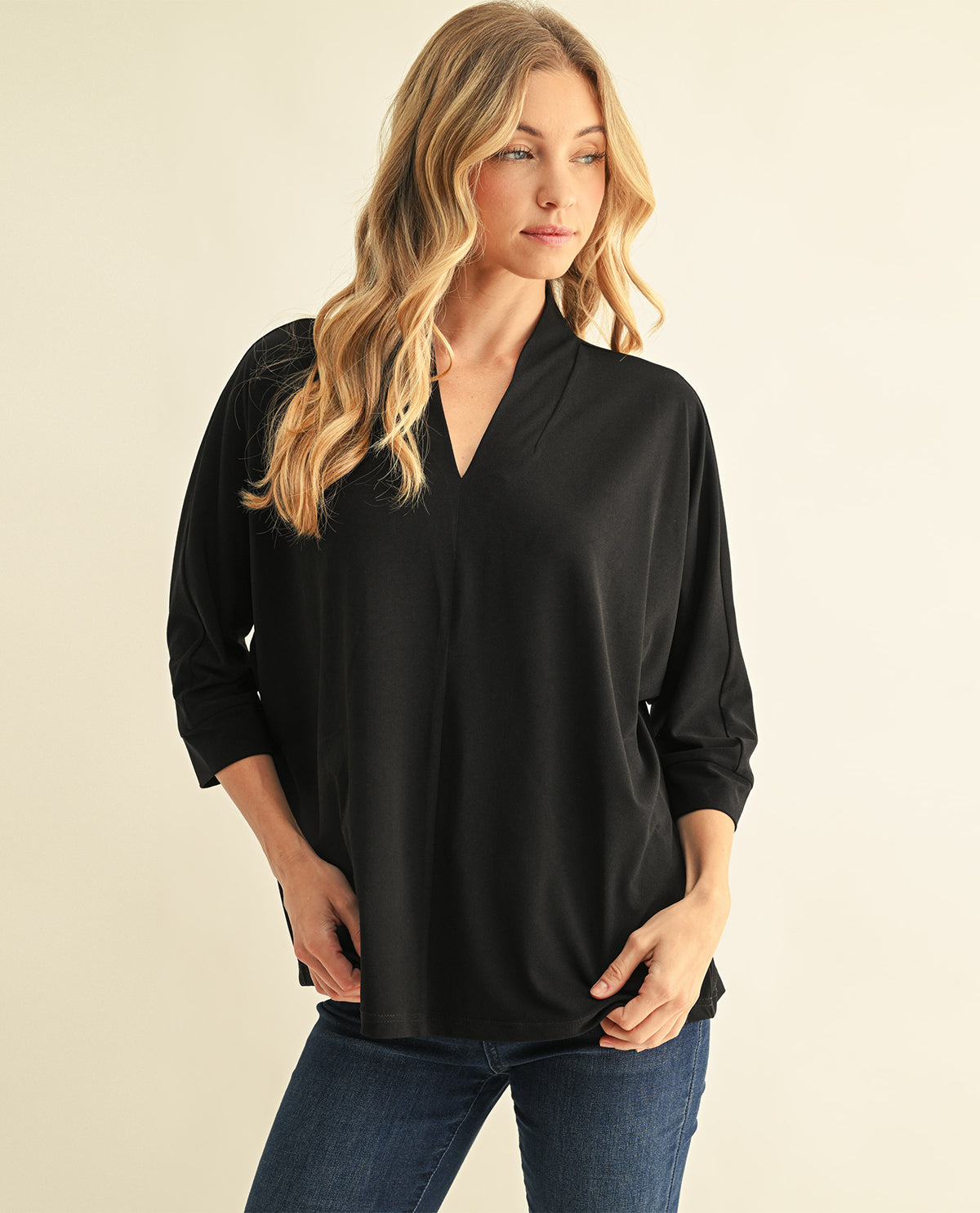 A woman modeling a black three-quarter sleeve top with a V-neckline, paired with blue jeans, against a solid light beige background.