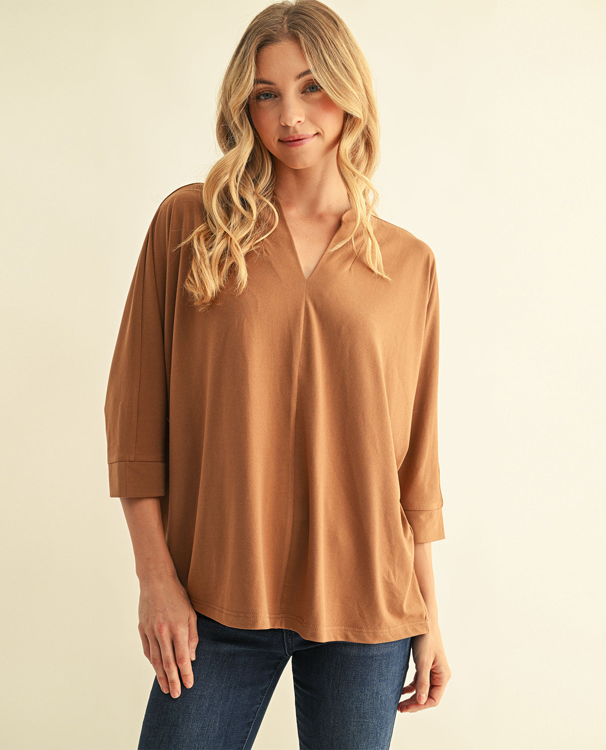A woman modeling a loose-fitting, three-quarter sleeve V-neck top in a warm camel brown color, paired with dark blue jeans, against a solid light beige background.