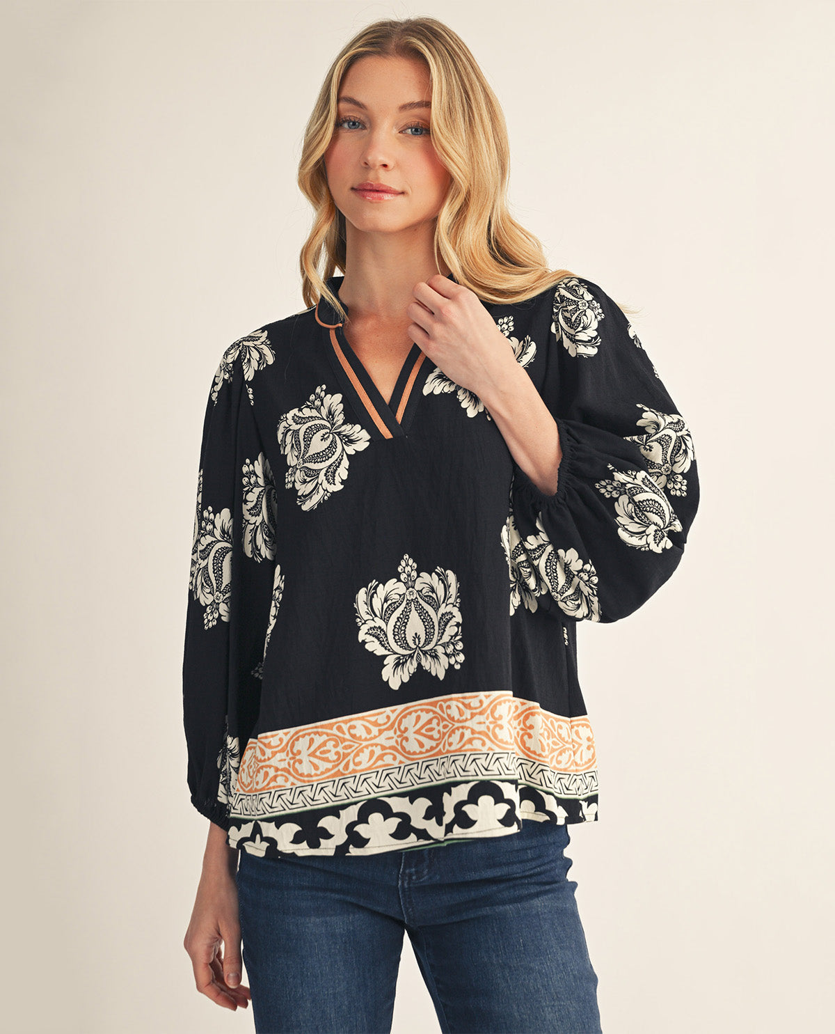 A woman modeling a black long-sleeve blouse with a white floral pattern and a peach and white border print near the hem, paired with blue jeans against a light beige studio background.