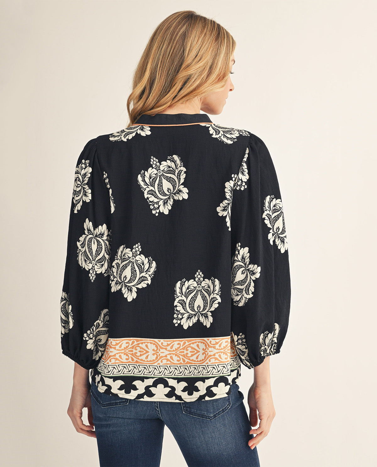A woman shown from the back wearing a black long-sleeve blouse with a white floral pattern and a peach and white border print at the hem, paired with blue jeans against a light beige background.