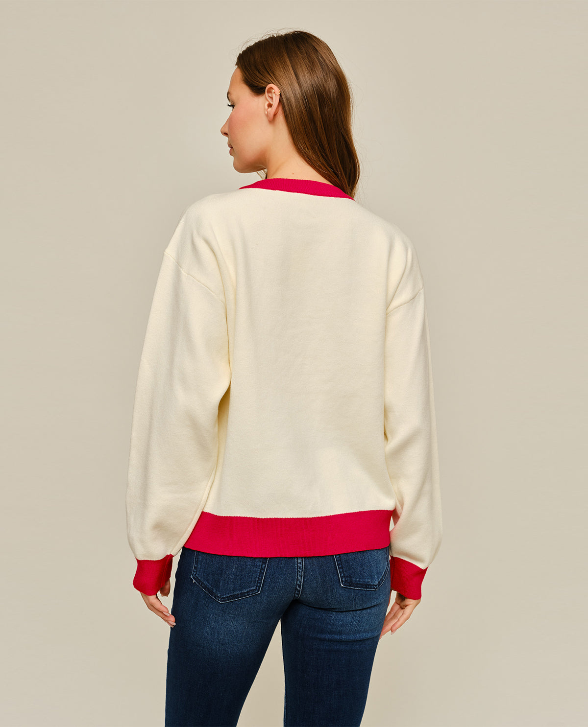 A woman shown from the back wearing a cream-colored crewneck sweatshirt with bright pink ribbed trim at the collar, cuffs, and hem, paired with blue jeans, against a solid light gray background.