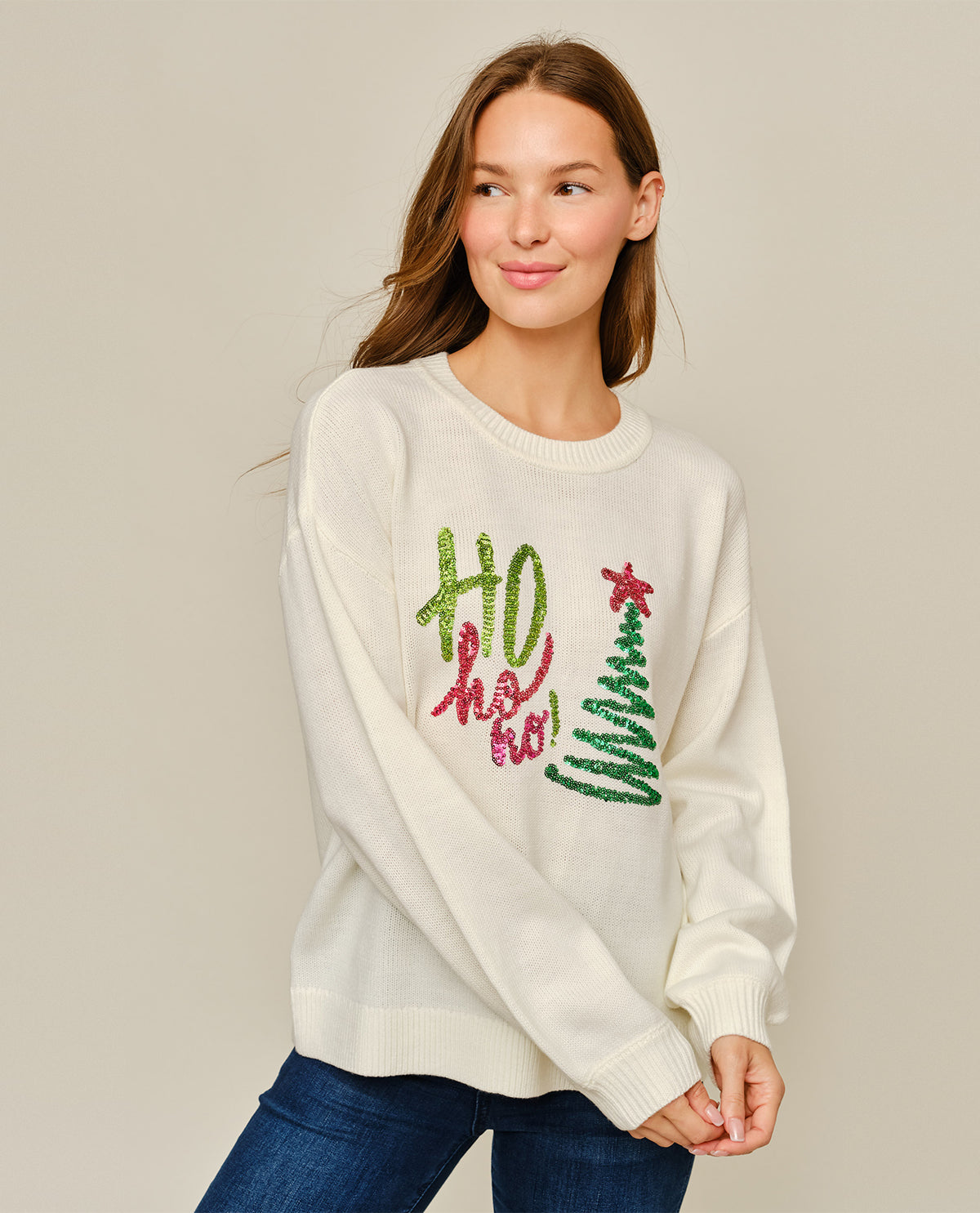 A woman wearing a cream-colored crewneck sweater with a festive 'Ho Ho Ho!' and Christmas tree design in green and pink sequins, paired with blue jeans, against a solid light gray background.