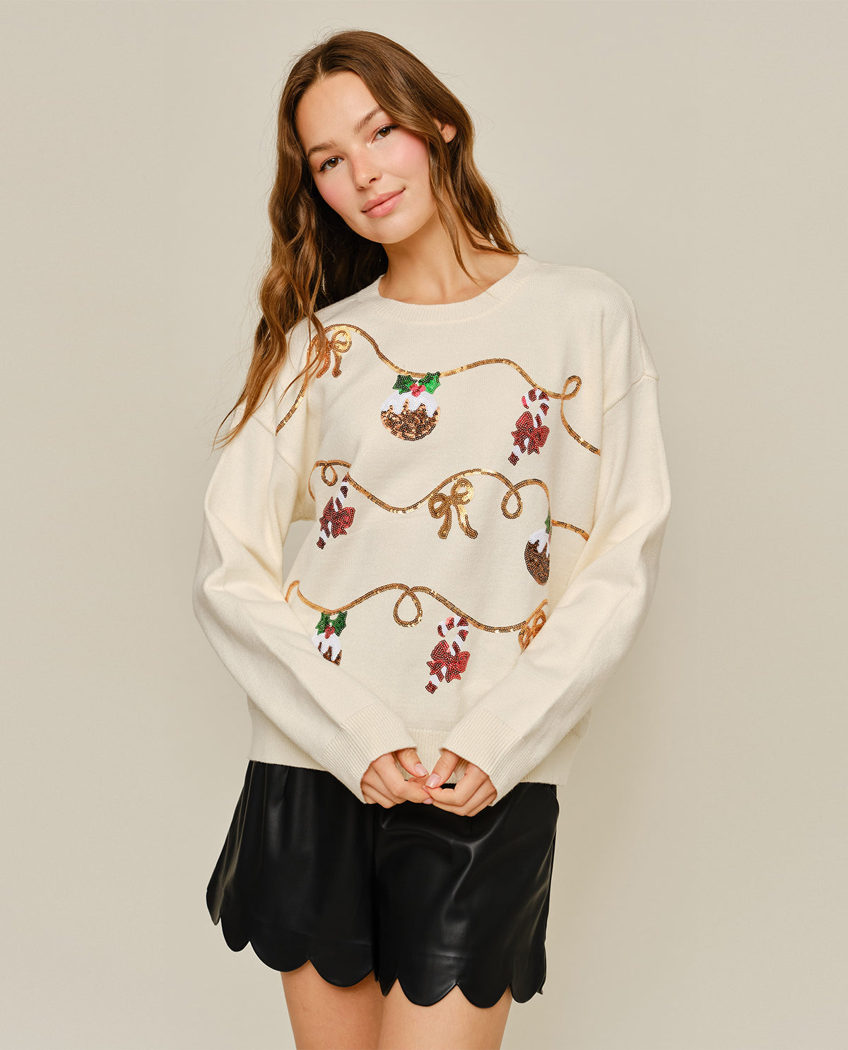 A woman modeling a cream-colored crewneck sweater with a brown bohemian-style floral and vine pattern, paired with black scalloped leather shorts, against a solid light beige background.