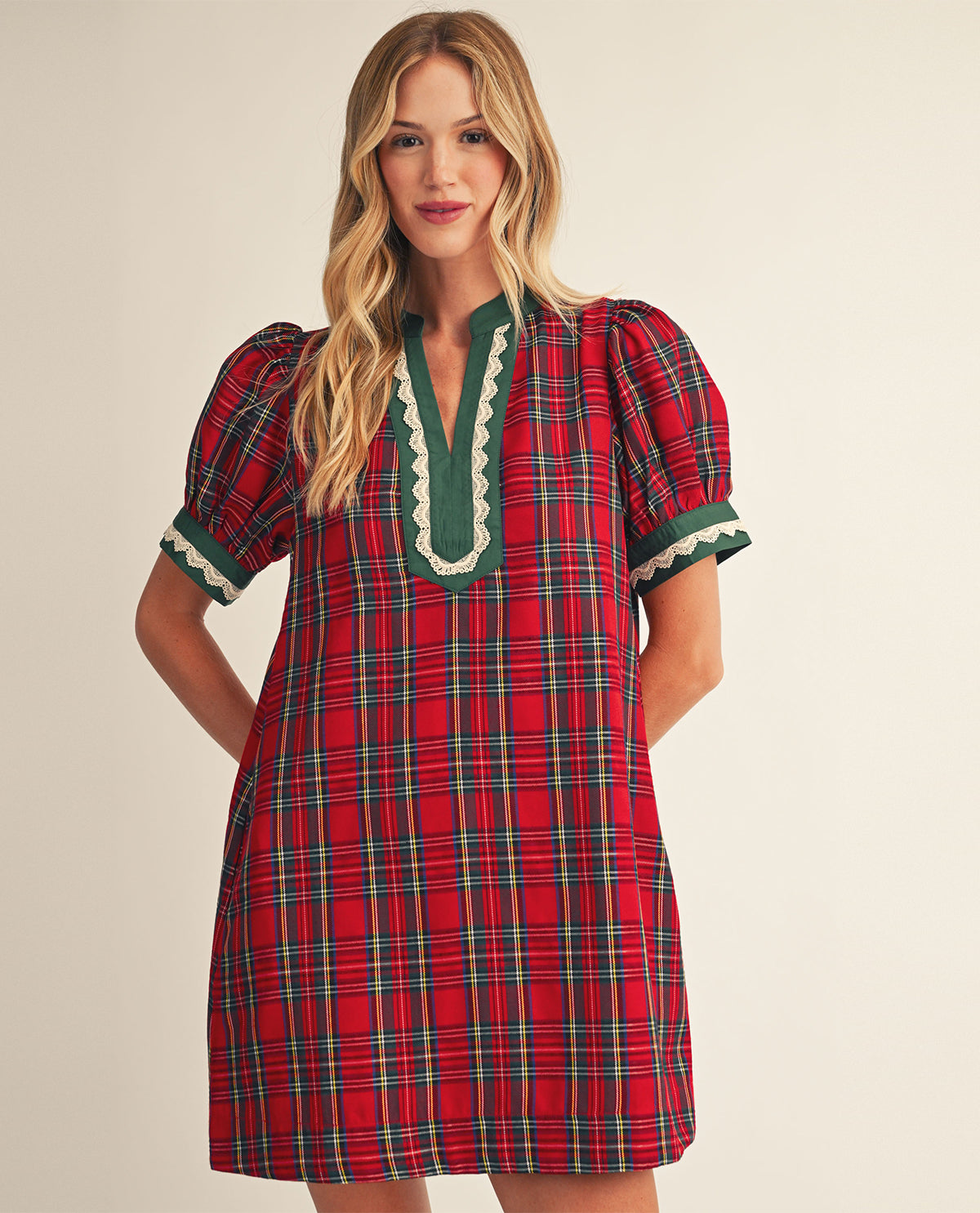 A woman modeling a red and green plaid short-sleeve nightgown with a green scalloped trim around the split neckline and cuffs, set against a plain beige studio background.