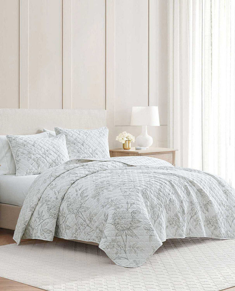A light gray and white botanical patterned quilt set on a bed in a bright, modern bedroom with a wooden nightstand, white lamp, and paneled wall.