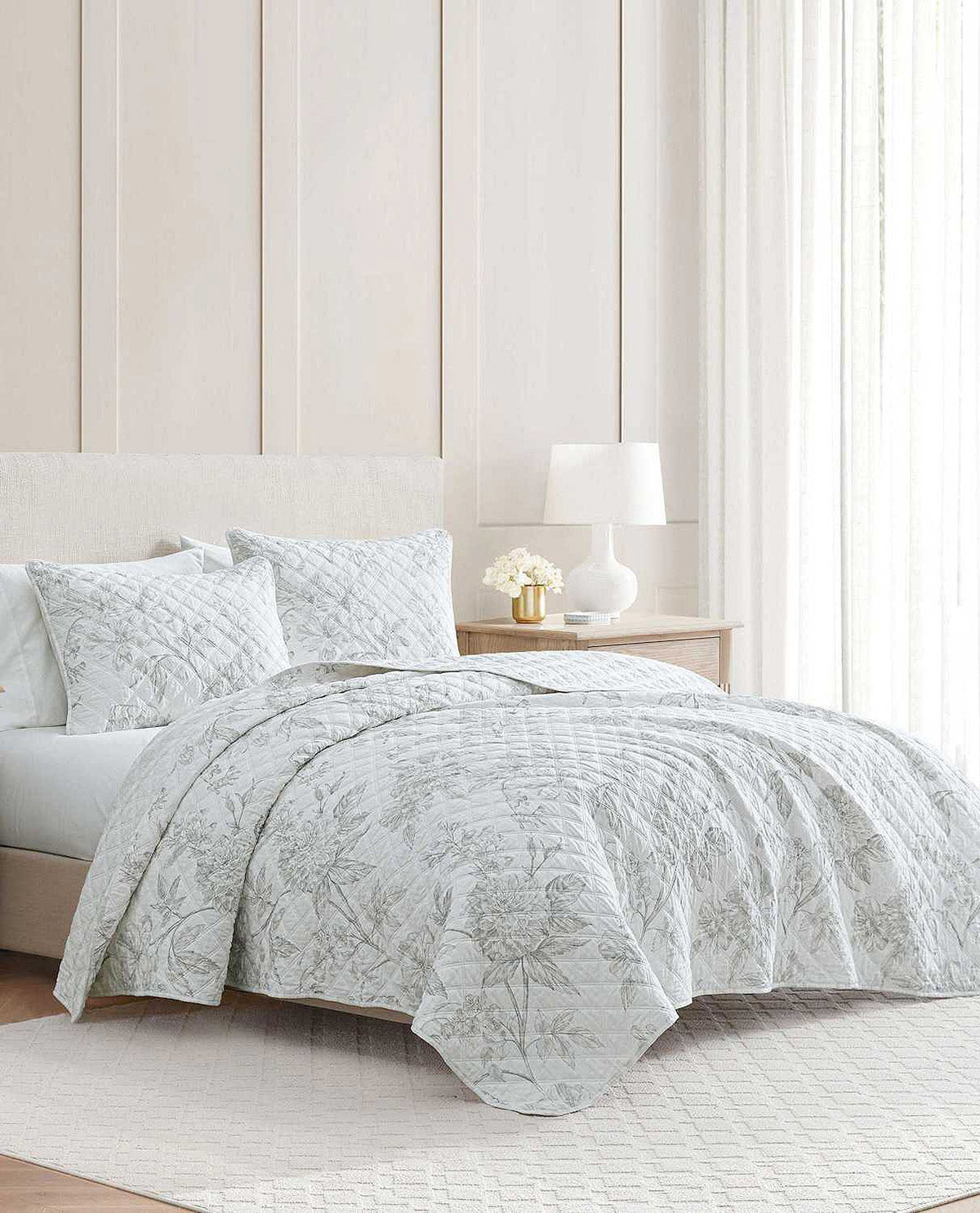 A light gray and white botanical patterned quilt set on a bed in a bright, modern bedroom with a wooden nightstand, white lamp, and paneled wall.
